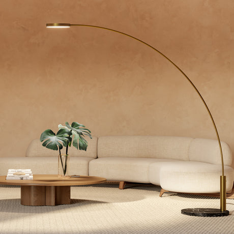 Dip Nero Marquina arc LED floor lamp with slanted brass arm, flat round head and black marble base, arching over a beige sofa and wooden coffee table.
