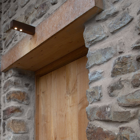 Zenthin W Wood LED outdoor wall light in Corten & Wood finish, slim rectangular bar with downward-facing LEDs, mounted beside a wooden door on a stone wall.