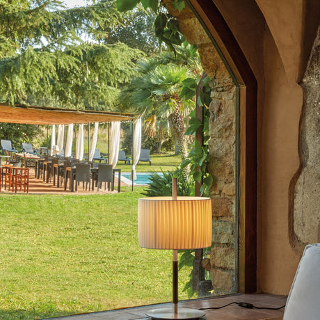 Danona Mini Ribbon Drum Table Lamp with pleated cream shade on a slim satin nickel stem and round base, shown by a window overlooking a garden terrace.