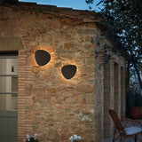 Two Tria 04 pebble-shaped outdoor wall lights in graphite brown metal, mounted on a stone wall, casting warm LED uplight and downlight glow.