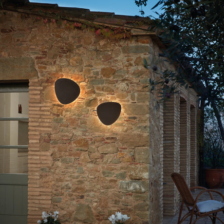 Two Tria 04 pebble-shaped outdoor wall lights in graphite brown metal, mounted on a stone wall, casting warm LED uplight and downlight glow.