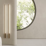Hilow Line linear wall lights in brushed bronze, two slim 150cm rods mounted on a curved beige wall beside a round window overlooking greenery.