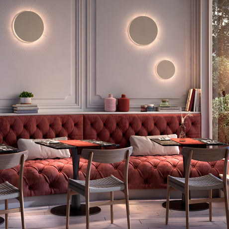 Round Helios A/01 LED wall lights with brown disc fronts and beige cotton thread edging, shown illuminated above a red tufted banquette in a dining area.