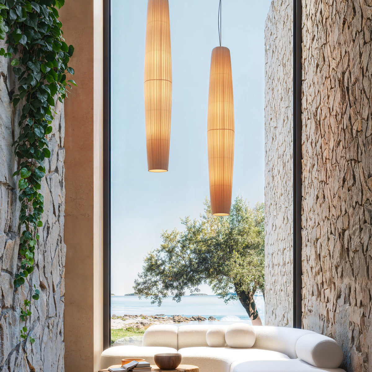 Maxi S/01 Ribbon LED pendant lights in cream, elongated pleated shades glowing warmly, hung beside a stone-walled window with sofa and sea view.