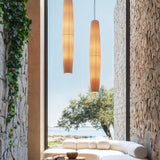 Maxi S/01 Ribbon LED pendant lights in cream, elongated pleated shades glowing warmly, hung beside a stone-walled window with sofa and sea view.