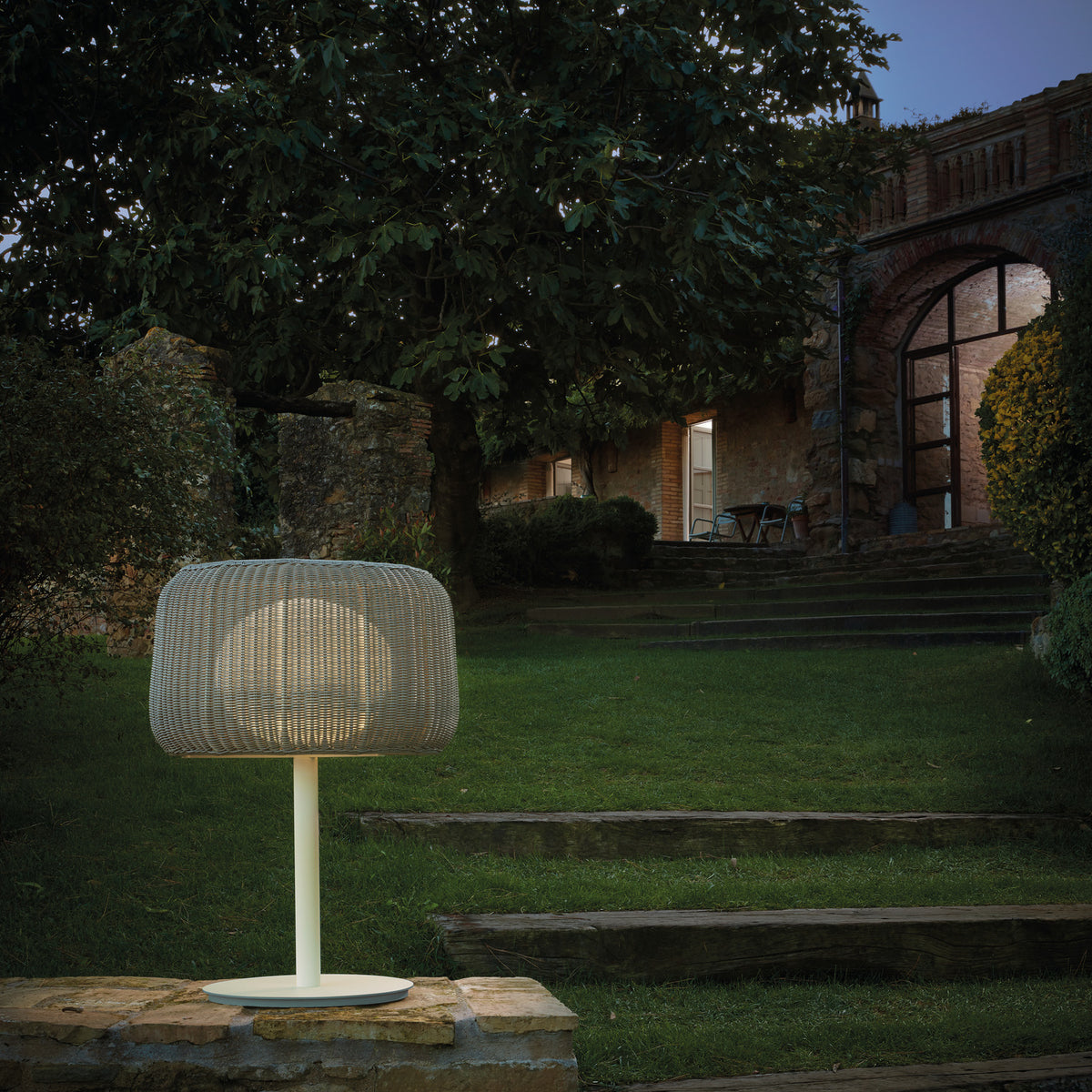 Fora M Drum LED Outdoor Table Lamp in Light Beige with woven drum shade, slim white stem and round base, glowing on a stone ledge in a garden setting at dusk.