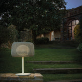 Fora M Drum LED Outdoor Table Lamp in Light Beige with woven drum shade, slim white stem and round base, glowing on a stone ledge in a garden setting at dusk.