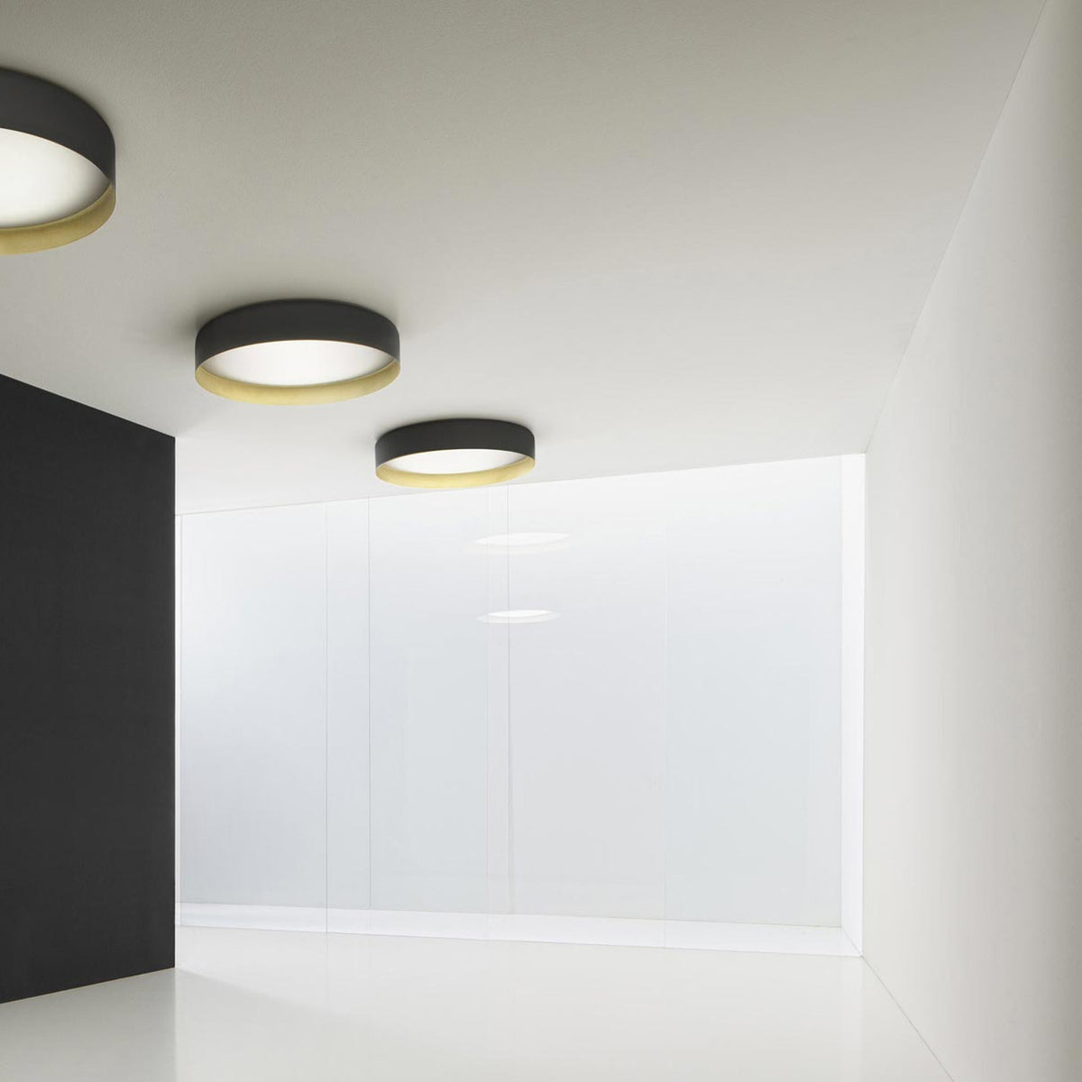 Ginevra 80cm round LED ceiling light in matt black with a brass inner band and white diffuser, shown mounted on a ceiling in a minimalist room setting.