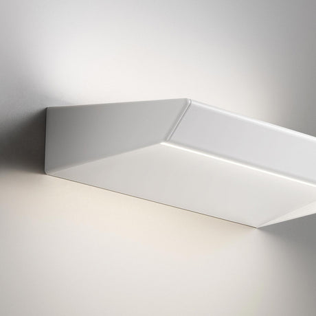 Close-up of the white Gonio LED Wall Light 44cm, showing its slim, folded wedge shape with angled edges and a hidden LED strip casting warm up-and-down light on a grey wall.