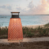 Nans Portable M/49/R rechargeable outdoor table lamp with red woven diamond-pattern shade, graphite cap and teak handle, glowing warmly on a beachside wall.