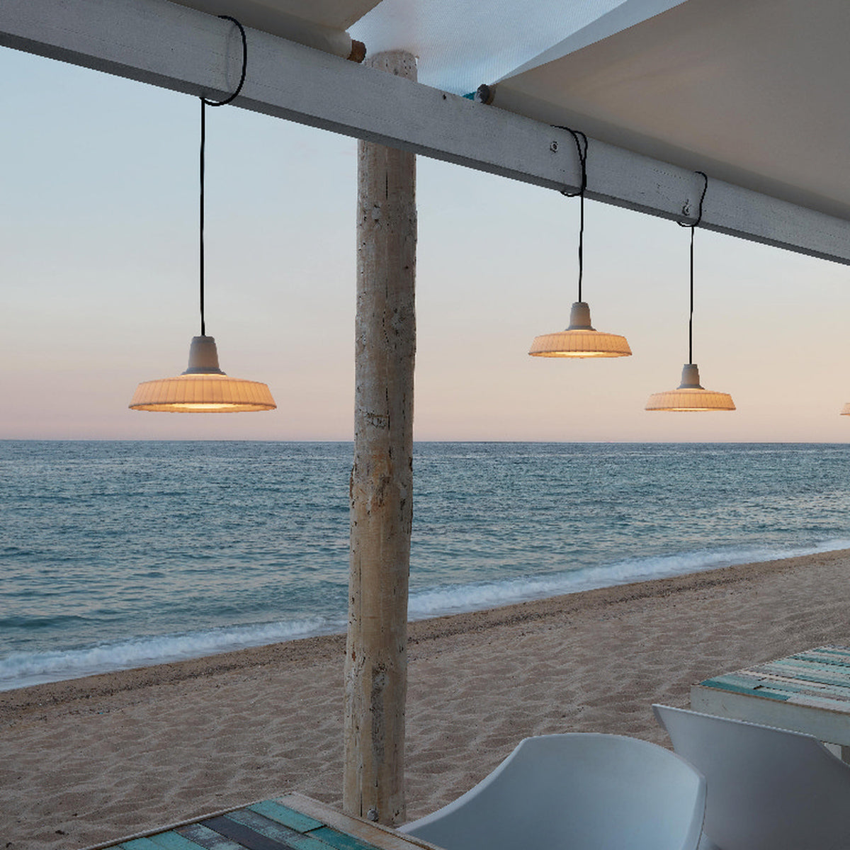 Marietta S/32 Dome LED Outdoor Pendant Light in white, with pleated dome shades on black cables, shown hanging from a beach pavilion beam against a sea and sky backdrop.