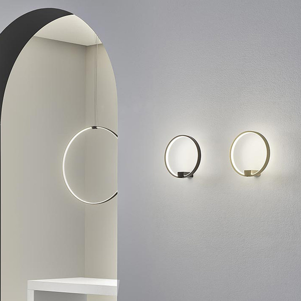 Zero Round Aluminium Ring LED Wall Light in black and brass finishes, shown illuminated on a grey wall beside a matching pendant version in an arched interior.