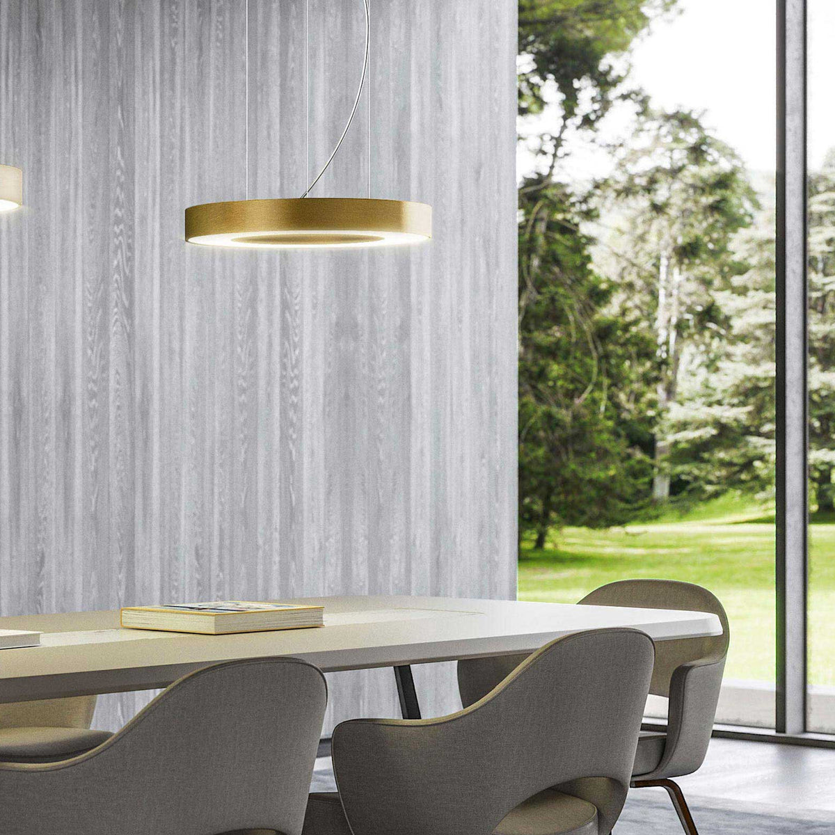 Silver Ring LED pendant light in gold leaf finish, a slim circular ring shade with integrated underside lighting, hung above a dining table in front of a grey wood-panelled wall and garden view.