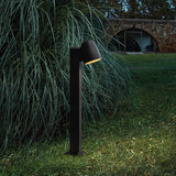 Nut B/60 Bell LED bollard light in textured black, slim square post with angled bell-shaped head, installed in a garden lawn beside tall grasses at dusk.