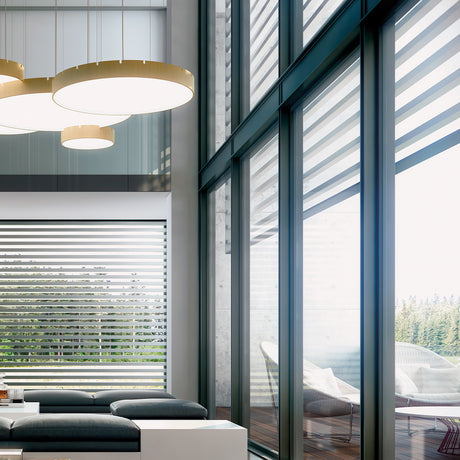 Castle S120 Aluminium LED Pendant Light in brass finish, shown as round drum-shaped fixtures with white diffusers, grouped above a living room seating area with floor-to-ceiling windows.
