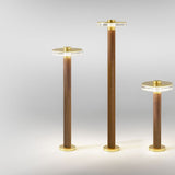 Venexia Glass LED Outdoor Floor Lamps in three heights, each with a slim wood column, round matt brass base and cap, and a clear glass disc head, shown lit on a plain light grey background.