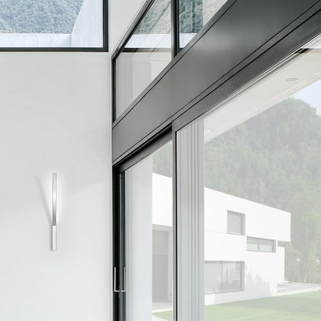 Ypsilon LED wall light in white, slim vertical bar fixture mounted beside a glazed sliding door in a modern interior with mountain views.