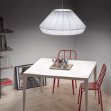Mei 125 Ribbon Cone pendant light in white, a wide pleated cone shade above a white dining table and red metal chairs.