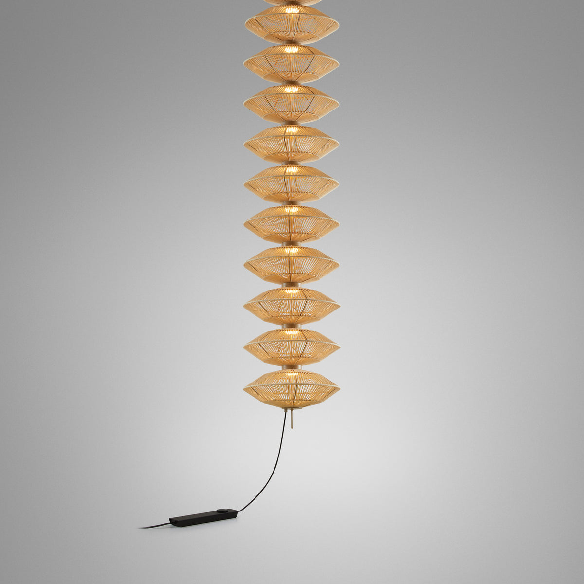 Yuyun S/11L/I pendant light with eleven stacked satin beige thread shades on a slim stem, illuminated, with black cord and foot dimmer, on a plain grey background.
