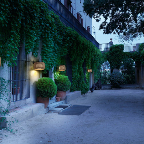 Fora A Drum LED Outdoor Wall Light in Light Beige, woven drum shade on a slim wall-mounted arm, shown illuminated along an ivy-covered building facade at dusk.