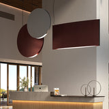 Noren S/100 fabric disc LED pendant light in Light Grey, a round panel with a glowing lower edge, shown alongside a rectangular burgundy pendant above a reception desk.