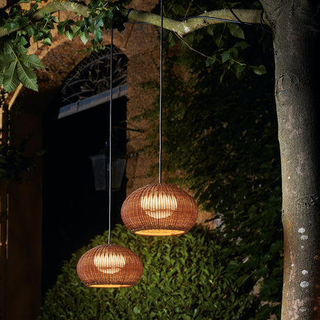 Garota Hang Globe LED outdoor pendant lights with brown woven open-weave shades, glowing warm white, hung from a tree branch near a garden doorway at night.