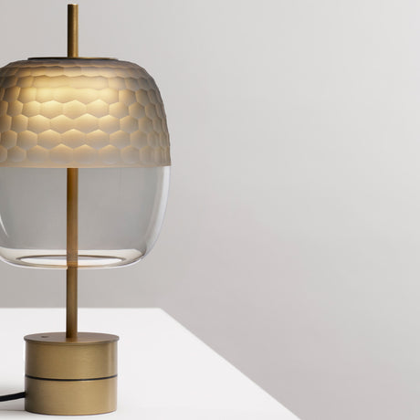 Adamas 23 table lamp with smoke grey glass shade, faceted honeycomb upper half, clear lower half, brushed brass stem and round base, on a light grey background.