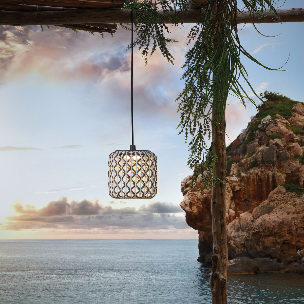 Nans S/16/H woven outdoor pendant light with beige barrel-shaped shade and integrated LED, hanging from a wooden beam against a coastal cliff and sky backdrop.