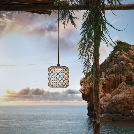 Nans S/16/H woven outdoor pendant light with beige barrel-shaped shade and integrated LED, hanging from a wooden beam against a coastal cliff and sky backdrop.