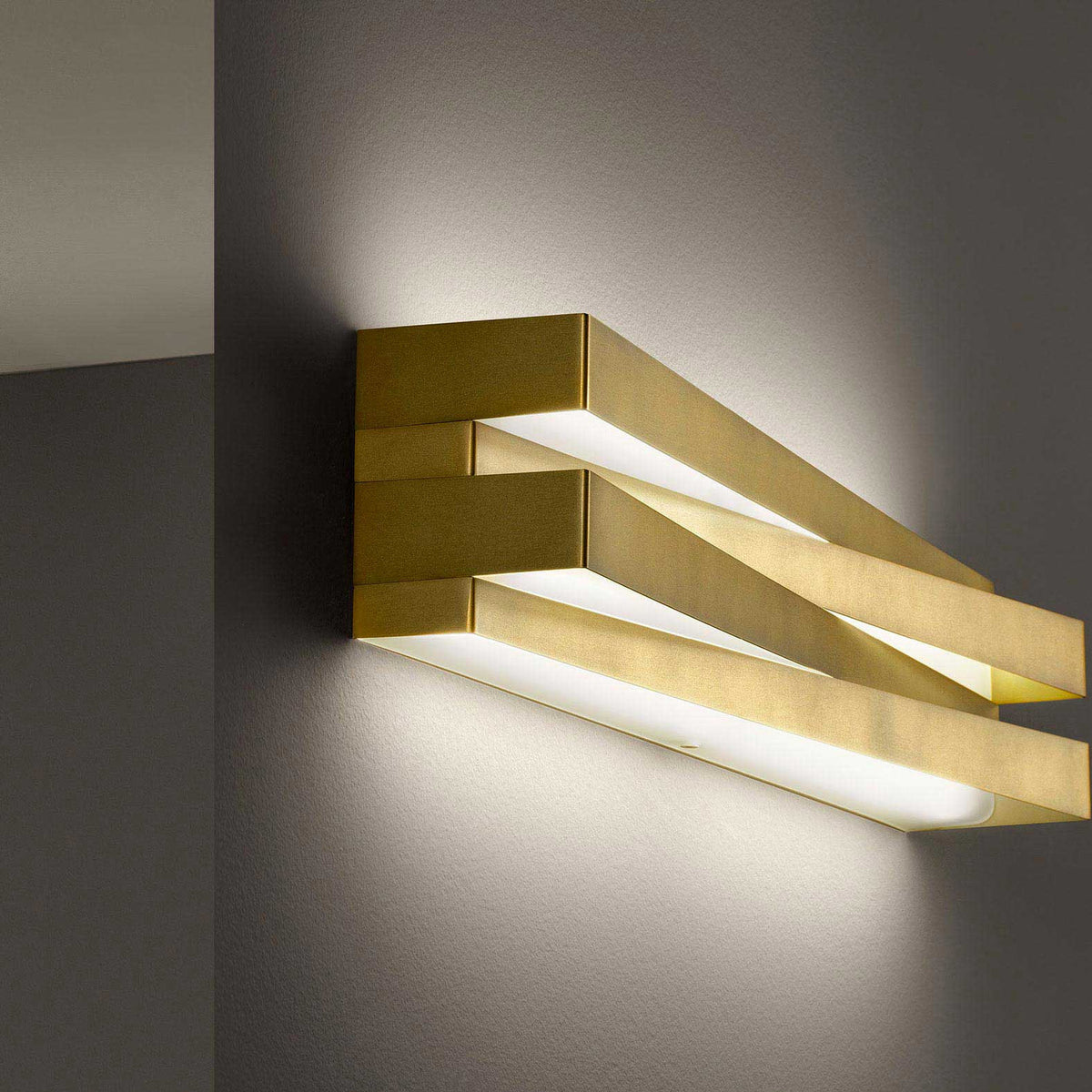 Cross Glass LED Wall Light 30cm in brushed brass, crossed bar design mounted on a grey wall, glowing warm white light along its length.