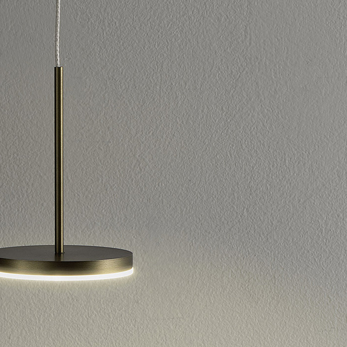 Bella Direct LED pendant light in brushed bronze with a slim round aluminium disc head and fine central stem, glowing warm white from its underside ring against a plain grey background.