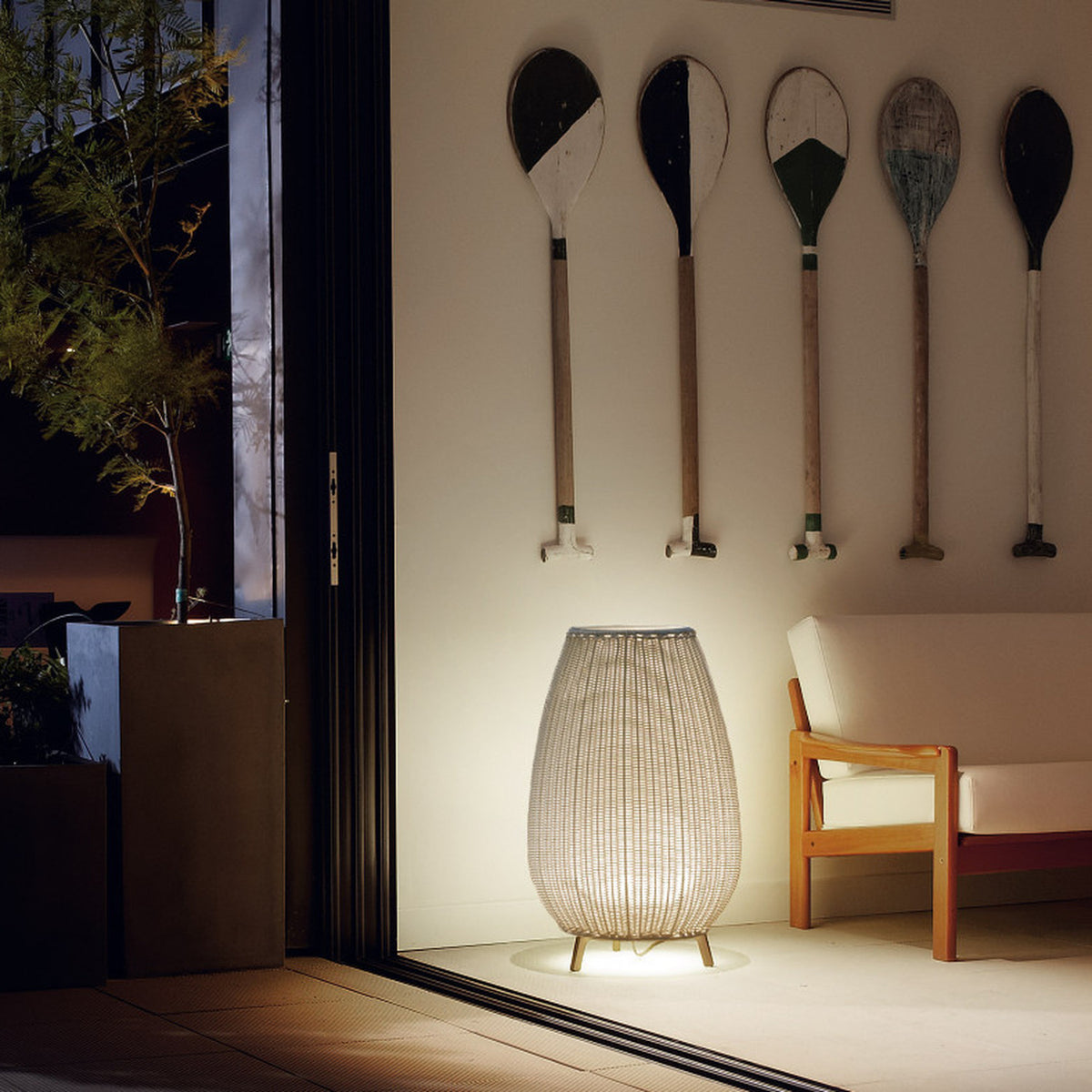 Amphora 01 beige woven LED outdoor floor lamp with tapered shade on three short legs, glowing beside a sofa under wall-mounted oars at night.