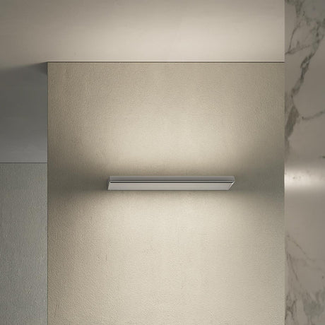 Gonio Linear LED Wall Light 58cm in Brushed Titanium, a slim wedge-shaped fixture mounted on a beige wall, casting warm uplight and downlight in a minimalist interior.
