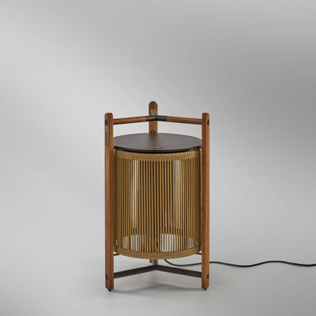 Kando P/76 outdoor floor lamp with sand nautical cord cylinder, three-legged teak wood frame, dark round lid, and wooden carry handle, shown on a plain light grey background.