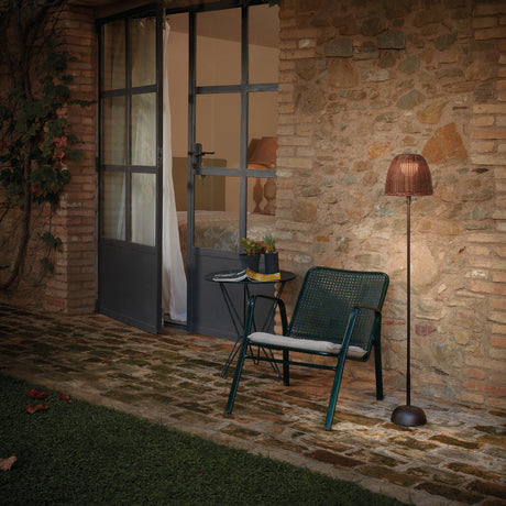 Atticus P/114 outdoor floor lamp with woven brown bell-shaped shade on a slim stem and domed base, lit beside a chair and side table on a stone patio at dusk.