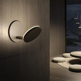 Bella Direct Aluminium Recessed LED Wall Light in bronze, round plate with angled disc head on a short stem, shown illuminated on a wood-panelled wall in a dimly lit lounge setting.