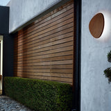 Tria 05 Wood LED Outdoor Wall Light in Teak Wood, a pebble-shaped fixture mounted on a textured grey wall, glowing softly against the surface beside a wood-slatted facade.