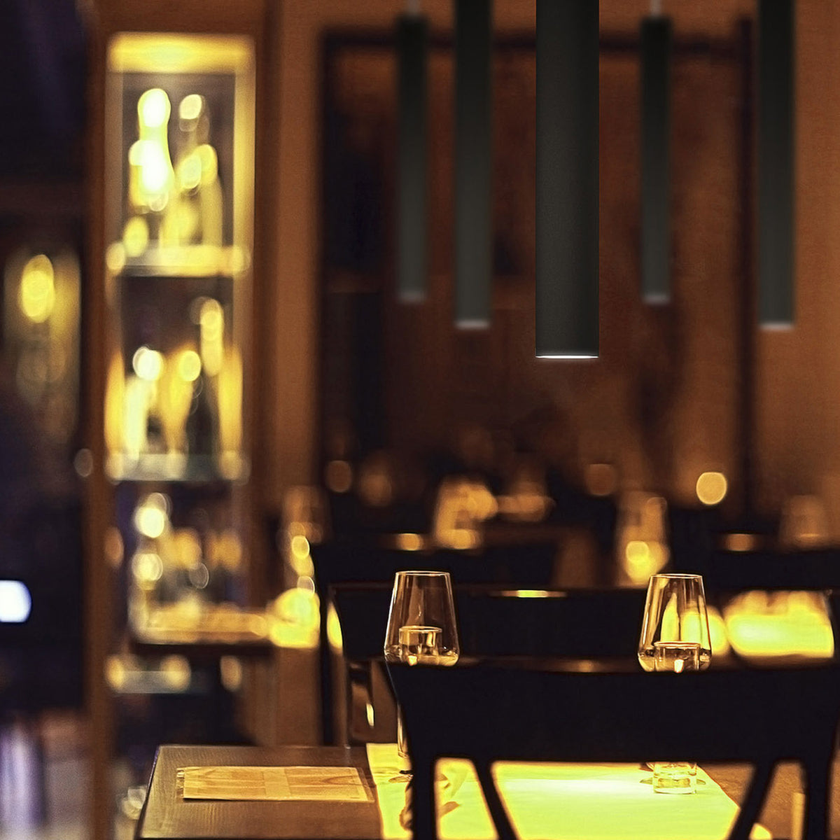 To-Be matt black aluminium tube LED pendant lights hanging above a restaurant dining table set with glassware, creating focused warm-white pools of light.