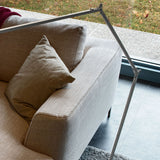 Jackie Aluminium LED floor lamp with jointed arm in brushed silver finish, shown angled above a beige sofa in a living room setting.