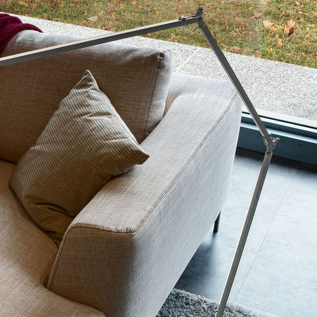 Jackie Aluminium LED floor lamp with jointed arm in brushed silver finish, shown angled above a beige sofa in a living room setting.