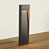 Zen Wood LED bollard light with a slim corten-framed column and dark wood front face, warm light glowing beneath the top head, shown outdoors on a wooden deck against a white wall.