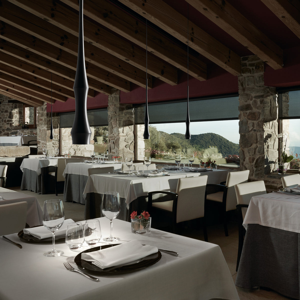 Slend 01 Aluminium Pendant Light in matte black with a slim, tapering tubular shade, shown hanging in multiples above a dining room with stone columns and timber beams.