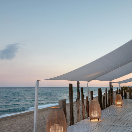 Amphora 02 Woven LED Outdoor Floor Lamps in beige, rounded fabric shades on short dark legs, illuminated along a wooden beach terrace deck at dusk.