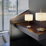 Danona T Ribbon Drum Table Lamps with pleated white shades on slim satin nickel stems and round disc bases, shown lit on a wooden shelf by a mesh-screened window.