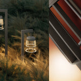 Coralys 120cm aluminium LED bollard light in Shadow Grey, shown outdoors among grasses, with slatted cage lantern glowing warm white beneath a curved arm.