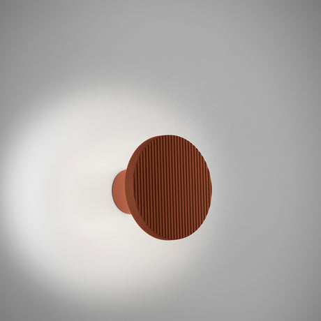 Arid A/01 round aluminium LED wall light in textured terracotta, ribbed disc on a short stem, casting a soft glow on a plain white wall.