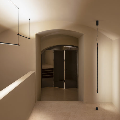 BIMU SV100 Metal Linear LED Pendant Light in black, mounted vertically from ceiling to floor anchor in a neutral hallway with arched doorway.