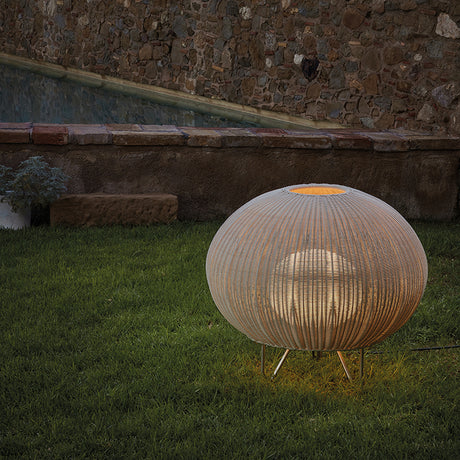 Garota P/01 Globe LED Outdoor Floor Lamp in Ivory White, a round woven globe shade on slim hairpin legs, glowing softly on a lawn beside a stone wall.