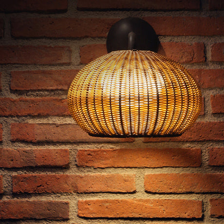 Garota A/01 outdoor wall light with a flattened globe woven brown shade on a curved arm from a round wall rose, mounted on a brick wall, integrated warm white LED.