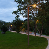 Zenete 300-2 Steel LED Bollard Light in brown finish, tall slim column with angled top arm casting warm white downlight along a garden path at dusk.